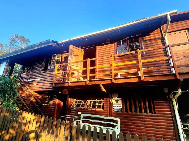A two-story timber log townhouse featuring an upper-level balcony, external staircase, and a picket fence in a natural setting.