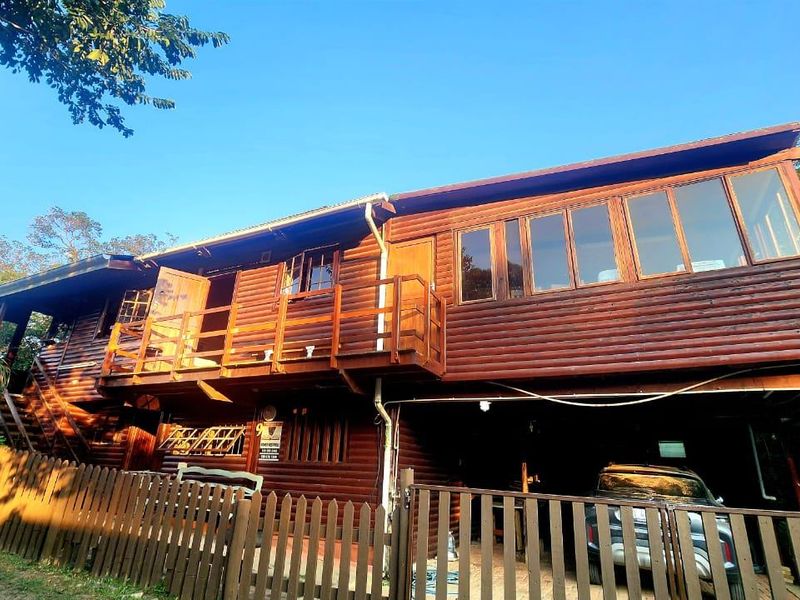 A two-story timber log townhouse featuring a raised balcony, external staircase, and a covered carport area, enclosed by a timber picket fence.