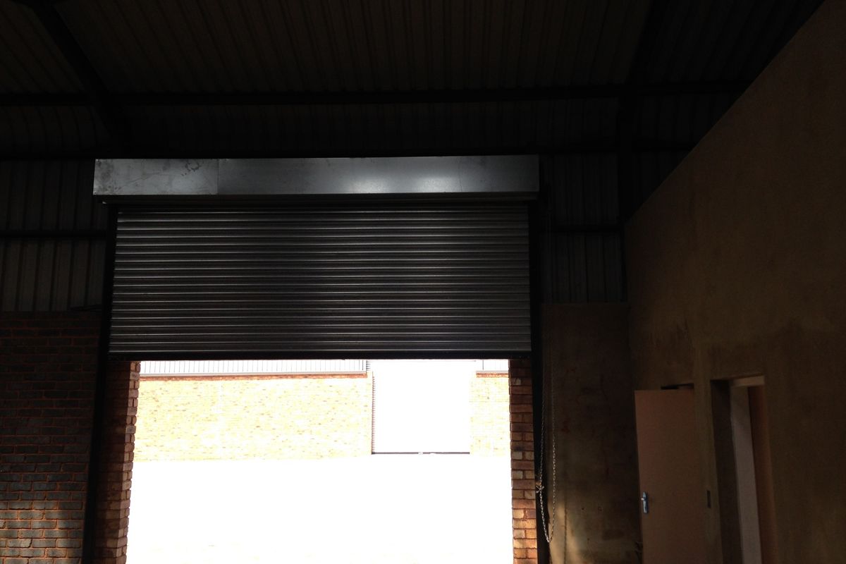 Industrial warehouse space featuring a large roller shutter door, exposed brick walls, and high-pitched metal roofing.