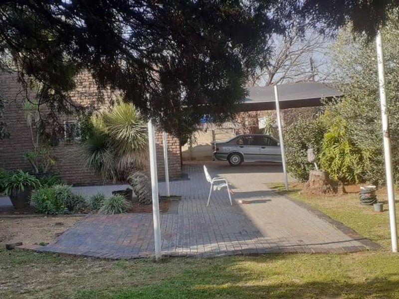A rear garden area featuring a paved patio, established landscaping, and a covered carport structure attached to the brick house.
