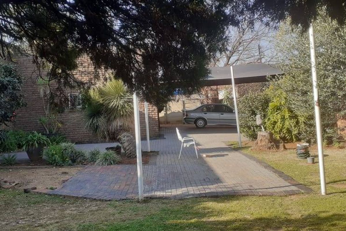 A rear garden area featuring a paved patio, established landscaping, and a covered carport structure attached to the brick house.