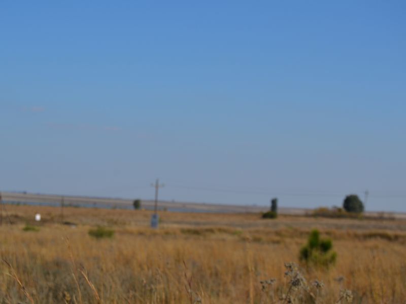 A wide-angle view of the vacant residential land, featuring open fields and distant utility infrastructure under a clear blue sky.