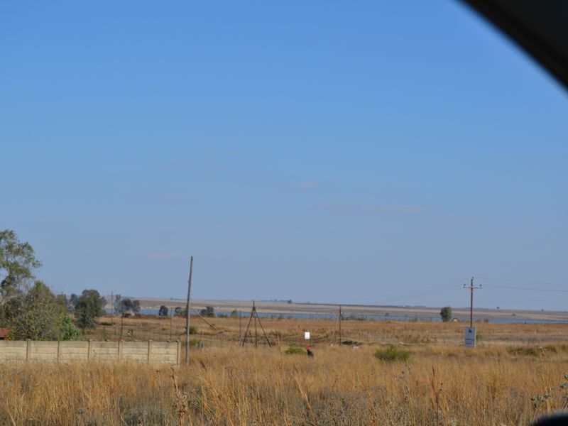 A wide-angle view of the vacant residential land, featuring dry grass, perimeter walling, and distant utility infrastructure under a clear blue sky.