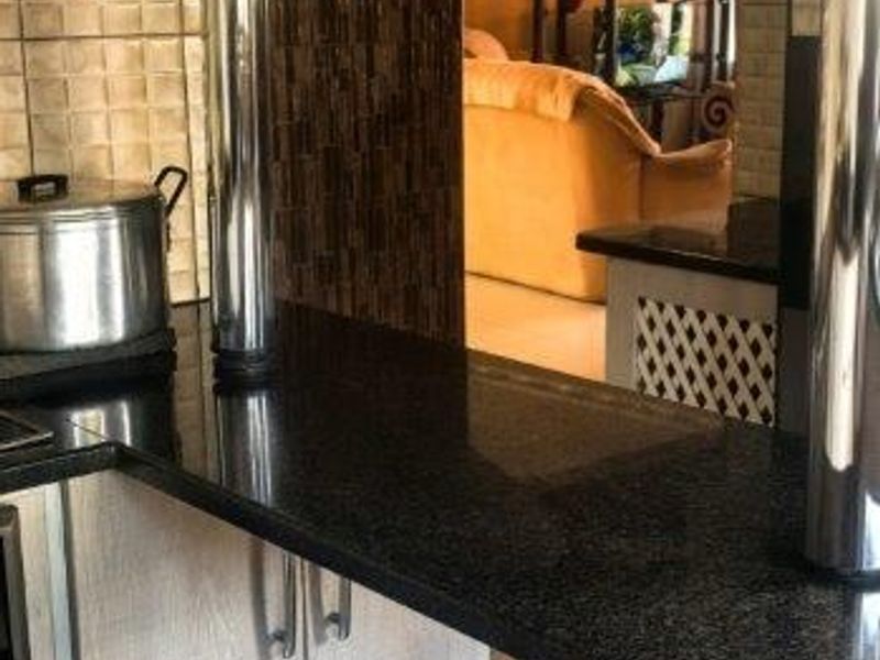 A kitchen area featuring granite countertops, light wood-finish cabinetry, and a tiled backsplash, with a breakfast bar overlooking the adjacent living space.