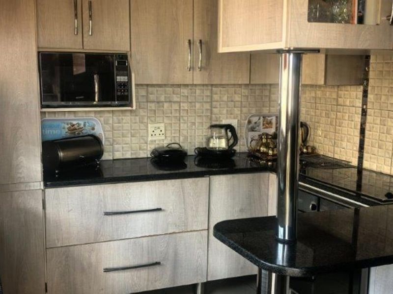 A functional kitchen featuring light wood-grain cabinetry, dark granite countertops, a tiled backsplash, and a convenient breakfast bar with a stainless steel support.