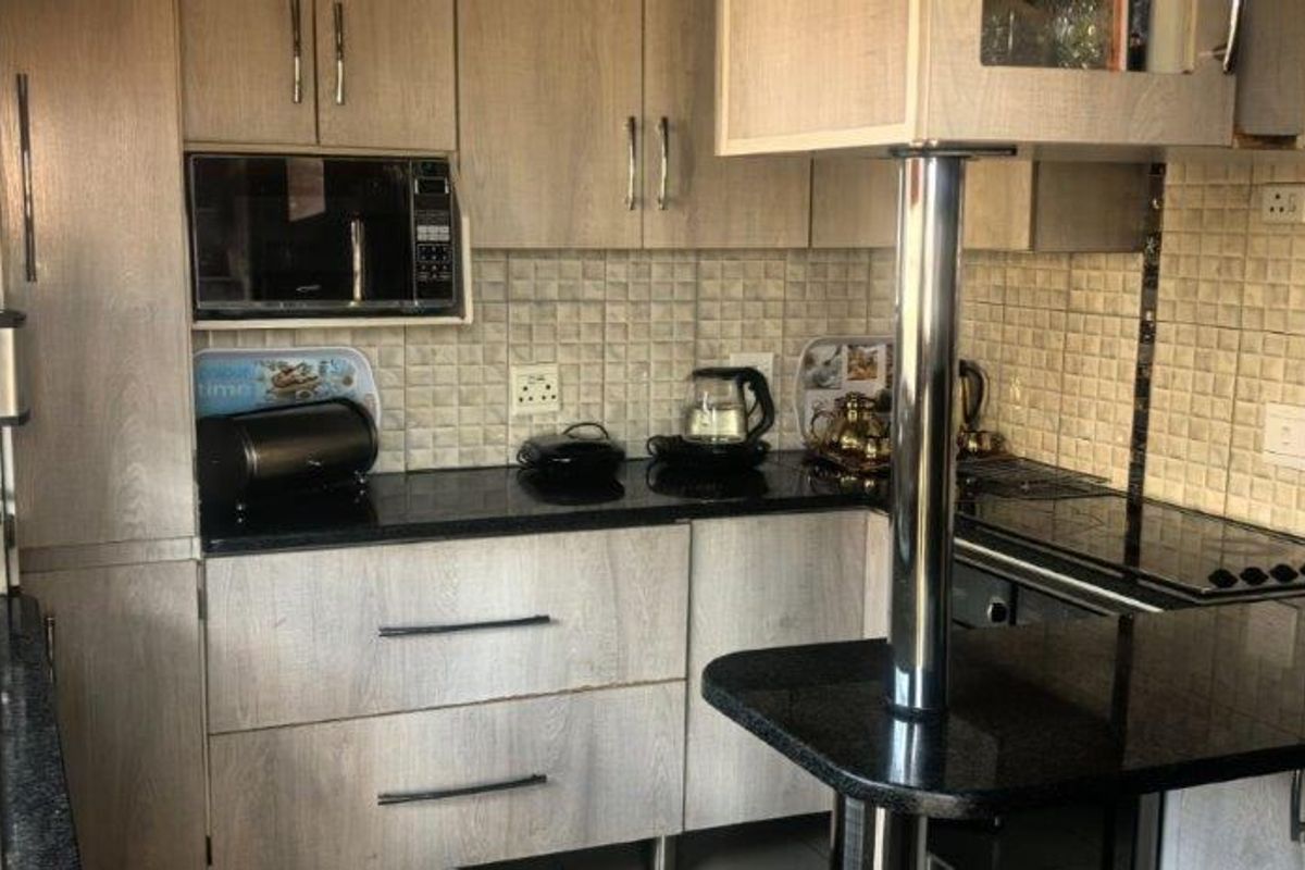 A functional kitchen featuring light wood-grain cabinetry, dark granite countertops, a tiled backsplash, and a convenient breakfast bar with a stainless steel support.