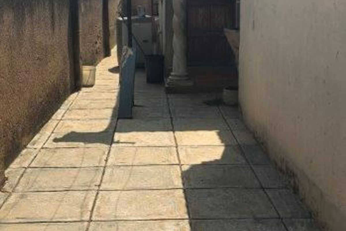 A narrow paved side passage or rear exterior walkway featuring a decorative column and boundary wall, requiring maintenance and surface cleaning.