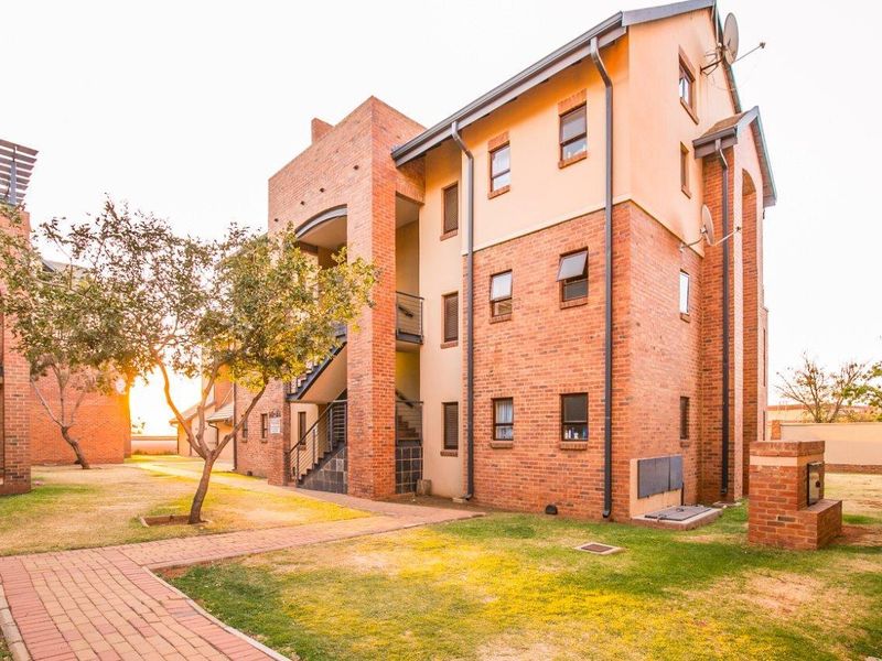 Exterior view of a multi-story face brick apartment building featuring paved walkways, external staircases, and a communal garden area with a built-in braai.