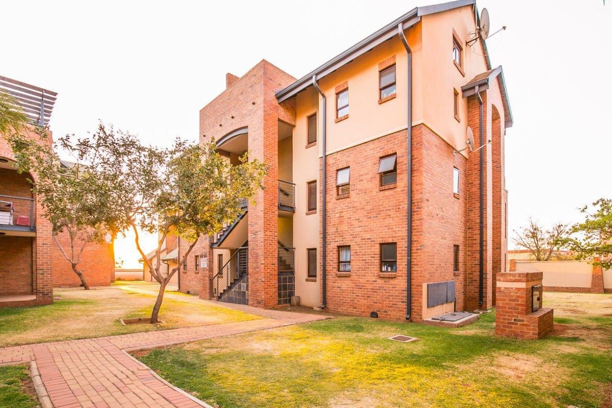 Exterior view of a multi-story face brick apartment building featuring paved walkways, external staircases, and a communal garden area with a built-in braai.