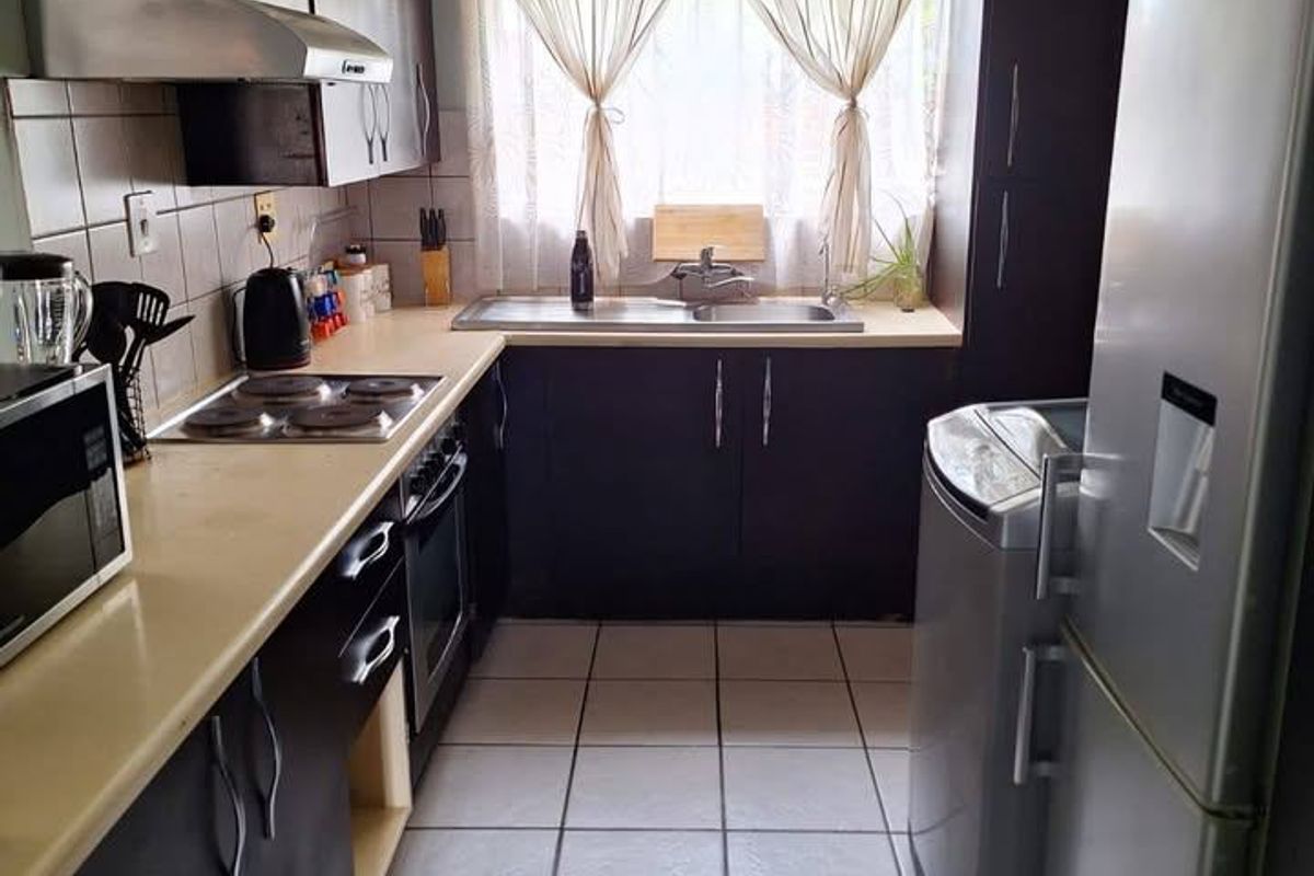 A functional galley-style kitchen featuring dark cabinetry, laminate countertops, built-in electric hob and oven, and tiled flooring with natural light from the window.
