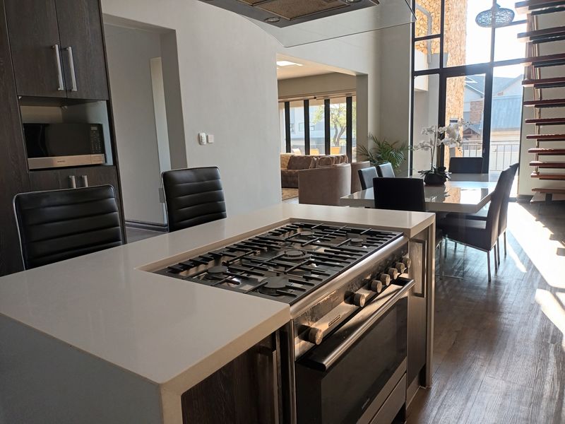Modern open-plan living area featuring a central kitchen island with gas cooktop, integrated appliances, and a striking timber staircase with floor-to-ceiling windows.