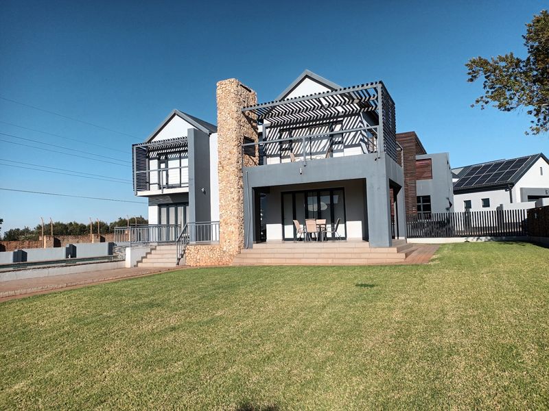 Rear view of a modern double-story house featuring a stone-clad chimney, upper-level balcony with timber pergola, and a spacious tiled patio overlooking the garden.