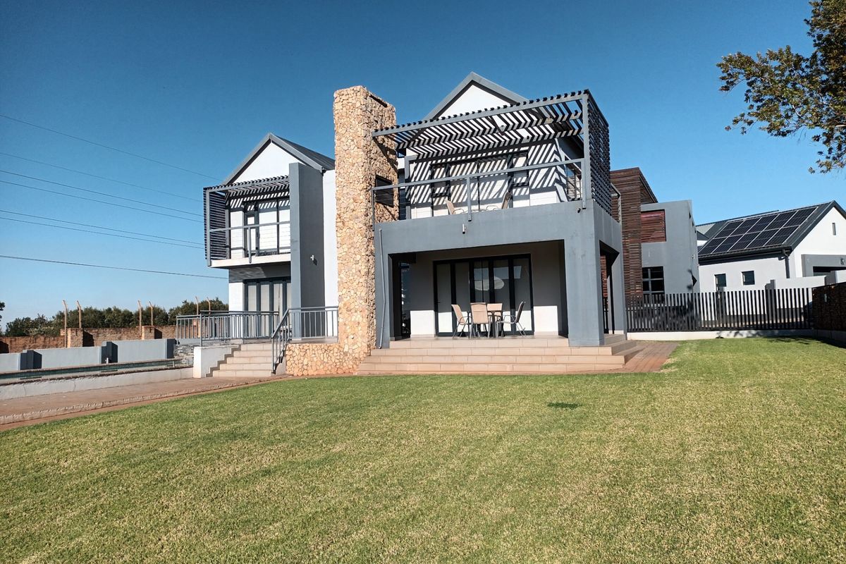 Rear view of a modern double-story house featuring a stone-clad chimney, upper-level balcony with timber pergola, and a spacious tiled patio overlooking the garden.