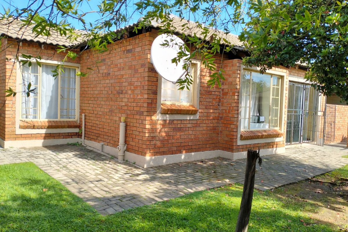 Rear view of the townhouse featuring a face brick facade, tiled roof, and a paved patio area with established garden surroundings.
