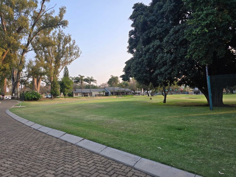 A well-maintained open green space featuring mature trees, a paved walkway, and established landscaping, providing a pleasant outdoor environment.
