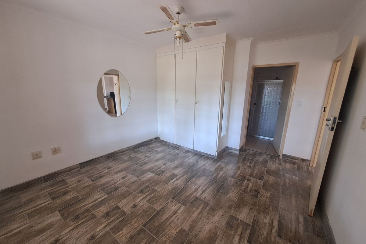 A bright, empty bedroom featuring wood-look tiled flooring, a built-in wardrobe, and a ceiling fan, with an adjacent en-suite bathroom visible.