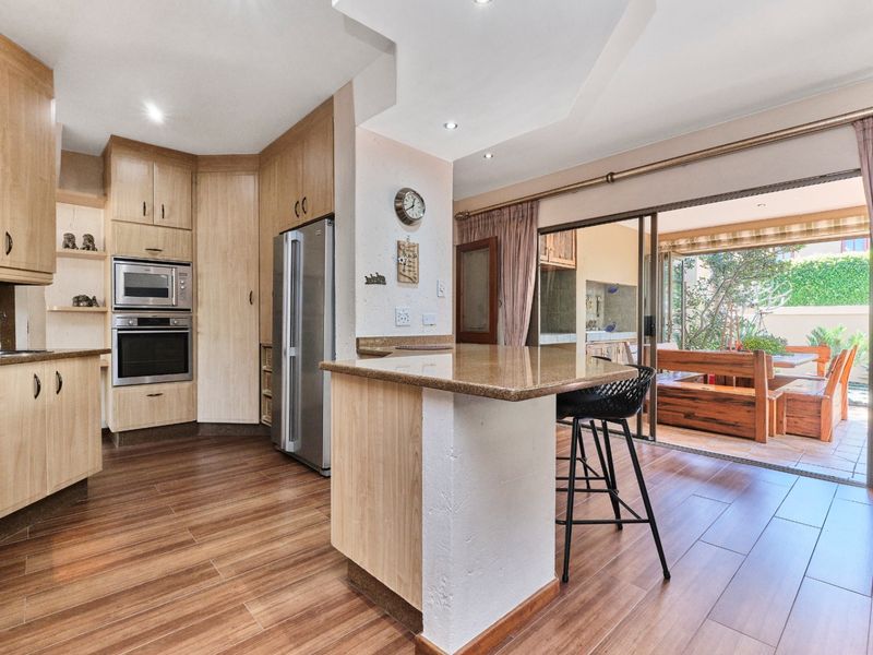 Open-plan kitchen featuring light wood cabinetry, granite countertops, and built-in appliances, flowing seamlessly to an outdoor patio area through large sliding glass doors.