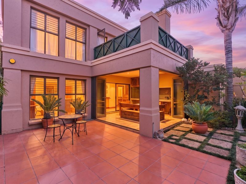 A well-maintained rear exterior view featuring a tiled patio, balcony, and large glass doors leading into the home, set against a landscaped garden.