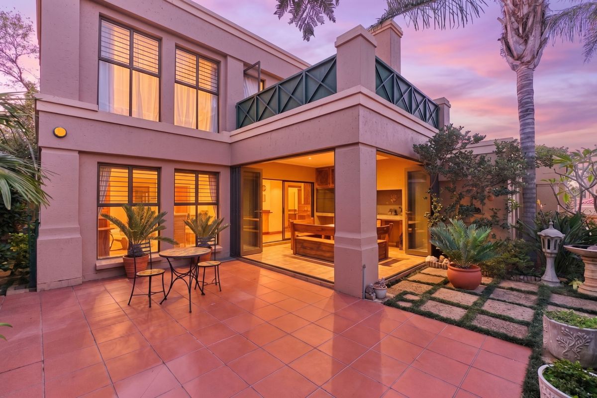 A well-maintained rear exterior view featuring a tiled patio, balcony, and large glass doors leading into the home, set against a landscaped garden.