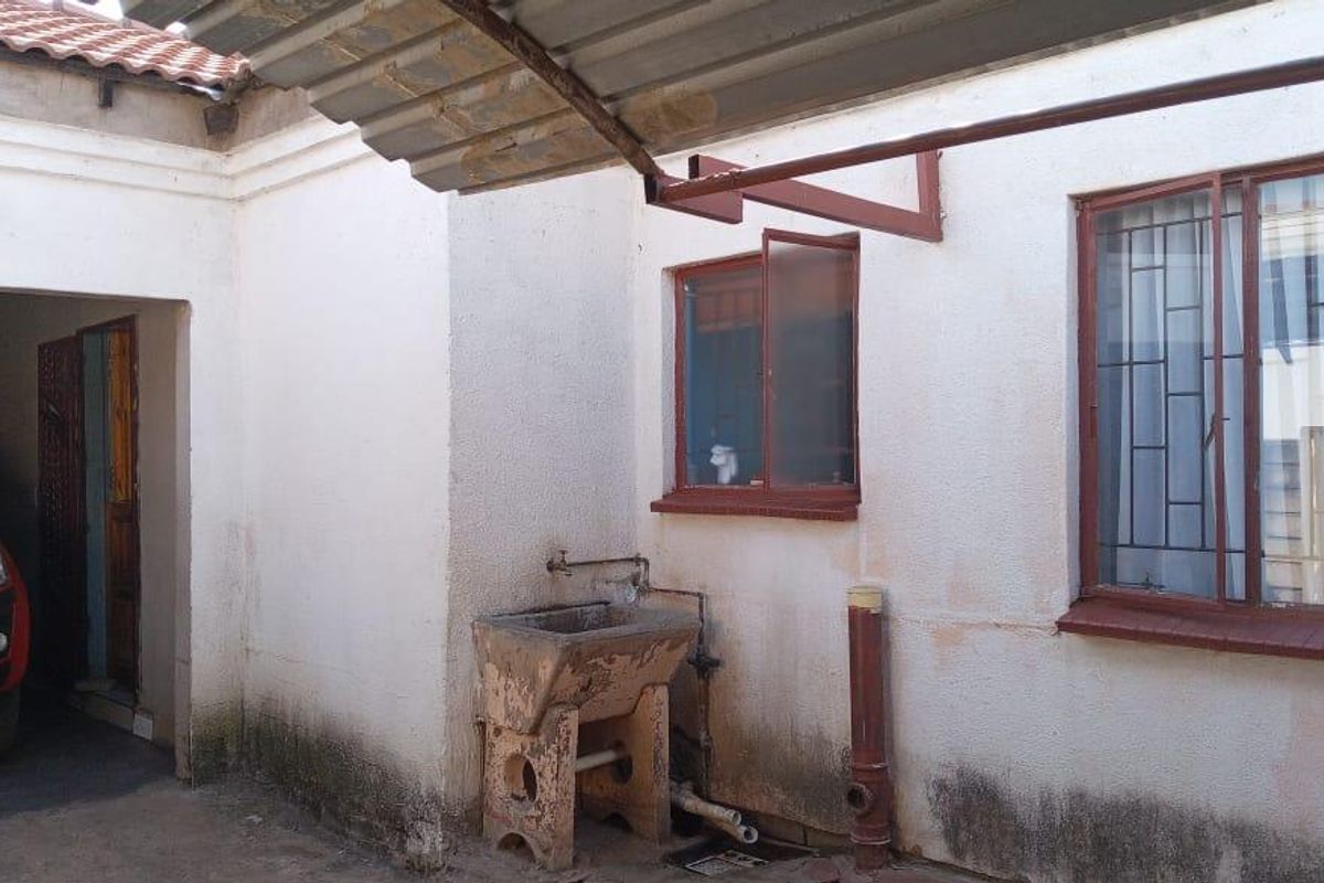 Rear exterior area featuring a concrete utility sink, metal awning, and security-barred windows, showing signs of significant wear and exterior wall staining.