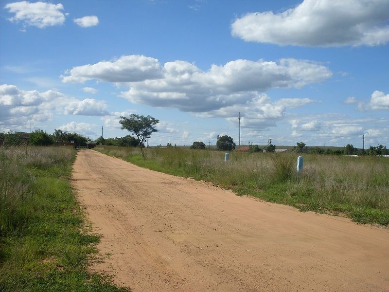 A view of an unpaved dirt road leading through a vacant residential plot with natural grass and boundary markers visible.