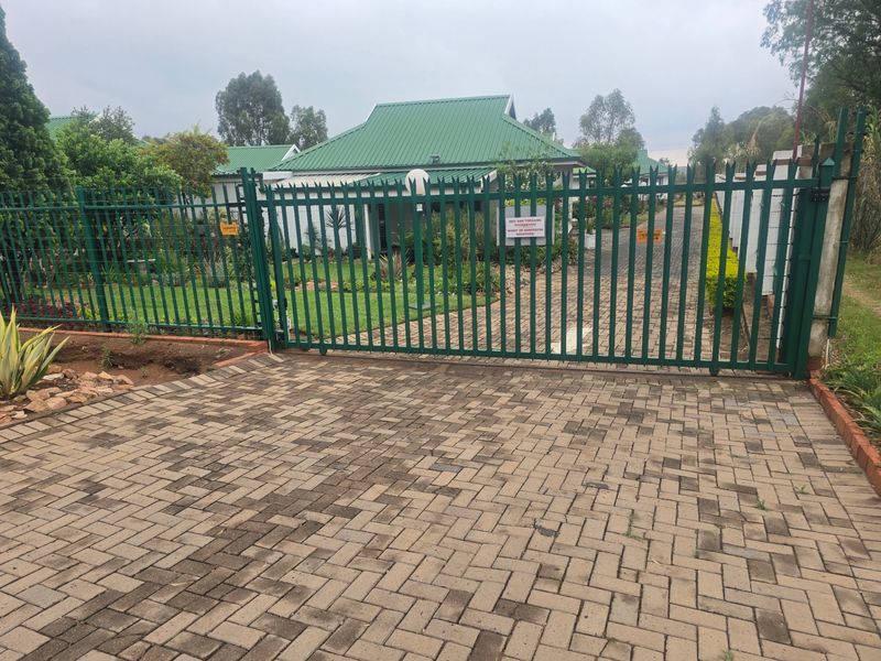Front view of a townhouse complex featuring a paved driveway, secure metal gate, perimeter fencing, and a well-maintained garden with a pitched metal roof.