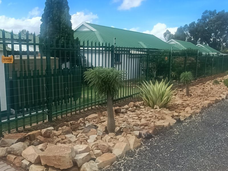Front exterior view of the townhouse complex featuring green metal security fencing, a paved driveway, and a landscaped rock garden.