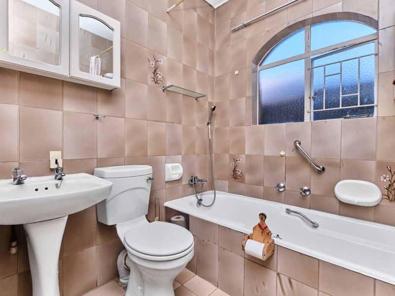 A dated bathroom featuring a pedestal basin, toilet, and bathtub with shower mixer, complemented by an arched window and wall-mounted storage.