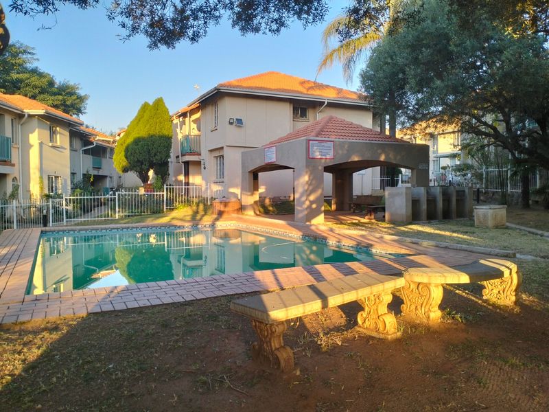 Communal pool area featuring a covered gazebo, built-in braai facilities, and stone seating, set within a well-maintained apartment complex garden.