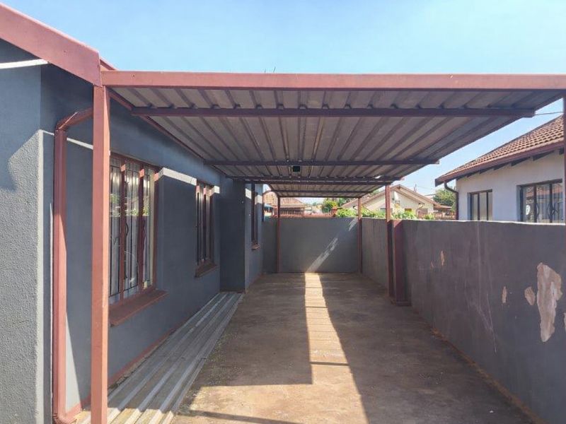 A covered carport area featuring a metal roof structure, concrete flooring, and a perimeter boundary wall with security-barred windows on the adjacent house wall.
