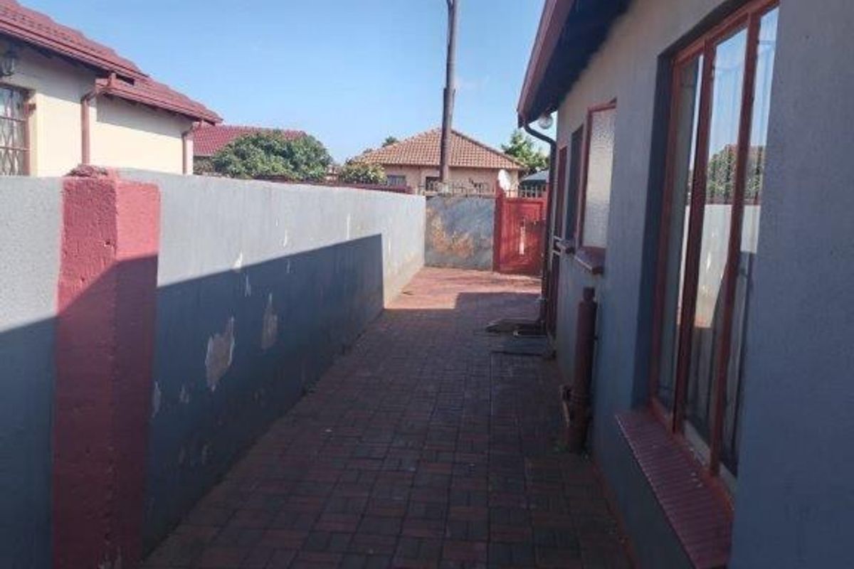 A narrow paved side passage along the exterior of the house, featuring a boundary wall and metal gate access.