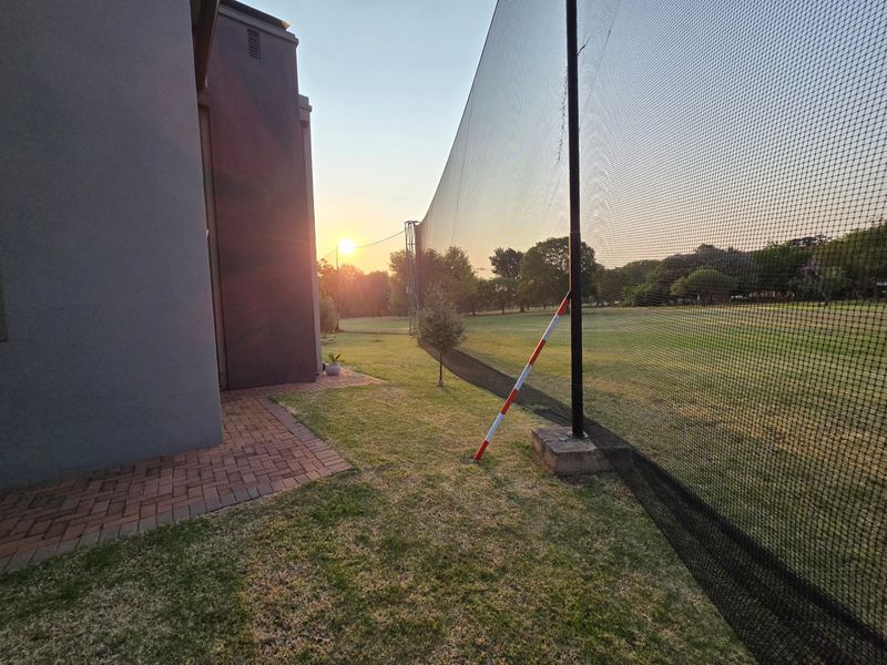 A rear garden area featuring a paved walkway alongside the house structure and tall sports netting bordering the lawn.