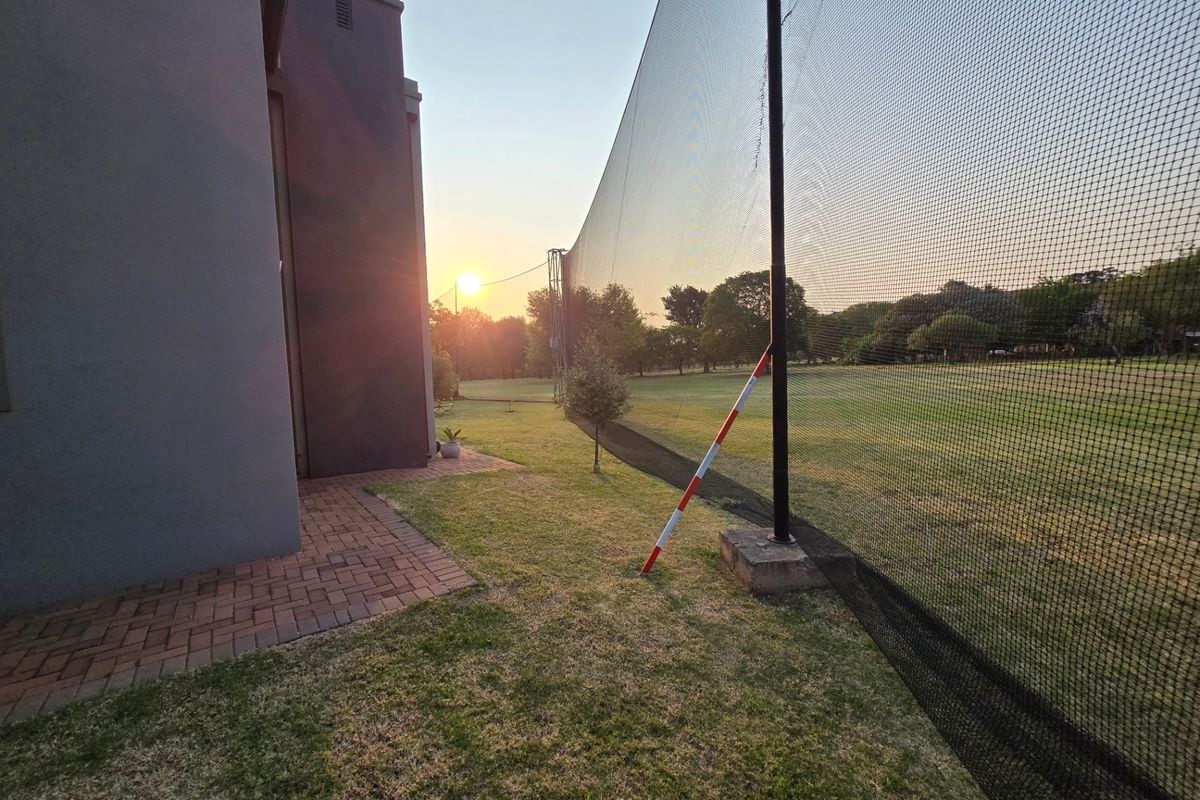 A rear garden area featuring a paved walkway alongside the house structure and tall sports netting bordering the lawn.