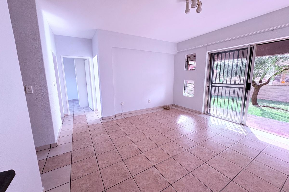 An empty, sunlit living room featuring neutral tiled flooring and a sliding glass door with security gate providing access to the garden.
