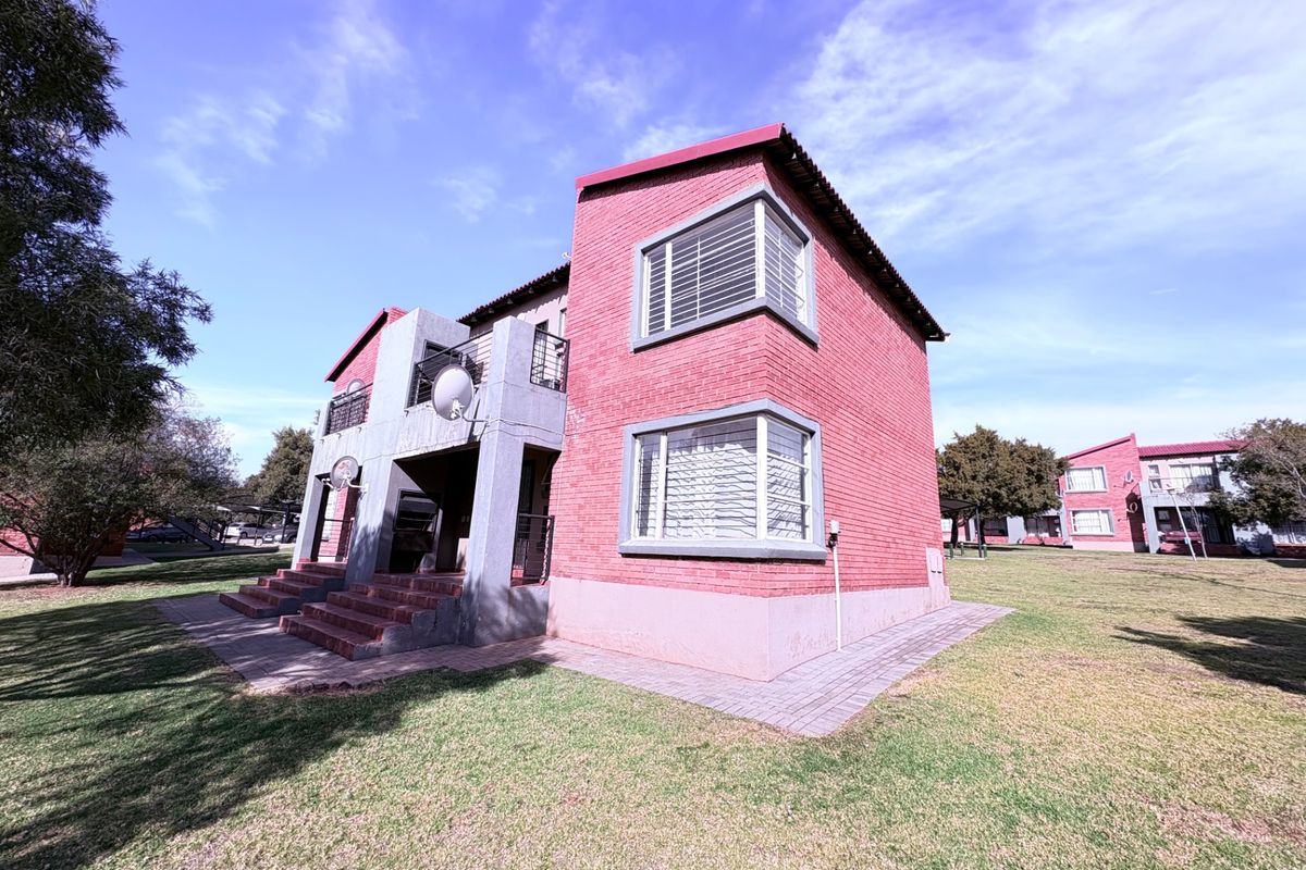 Exterior view of a face brick apartment building featuring balconies, metal security bars, and surrounding lawn area.