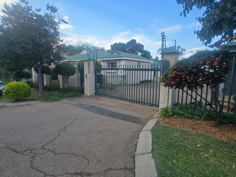Front view of the townhouse property featuring a secure metal gate, electric fencing, and a paved driveway with established garden landscaping.