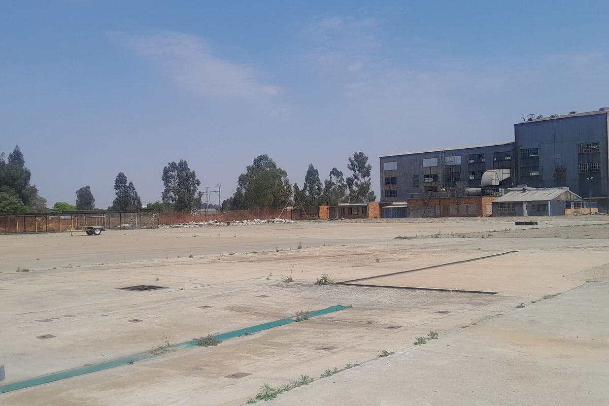 A large, vacant industrial concrete yard featuring perimeter fencing and an adjacent industrial building structure.