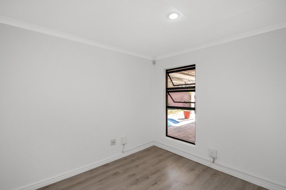 A clean, empty bedroom featuring modern laminate flooring, neutral white walls, and a window overlooking the outdoor area.