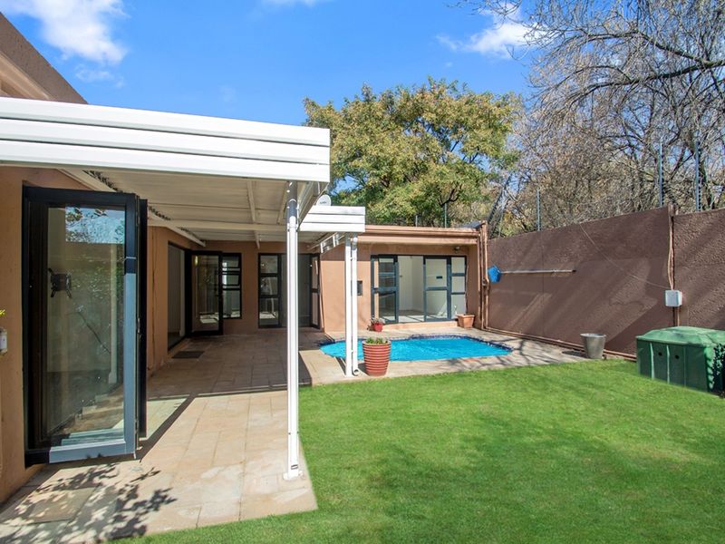 Rear exterior view featuring a covered patio, paved entertainment area, swimming pool, and a well-maintained lawn enclosed by a boundary wall.