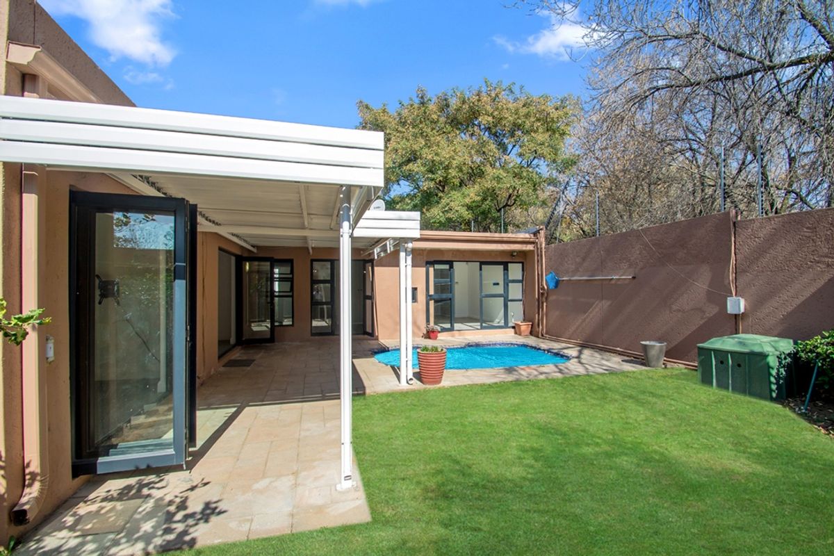 Rear exterior view featuring a covered patio, paved entertainment area, swimming pool, and a well-maintained lawn enclosed by a boundary wall.
