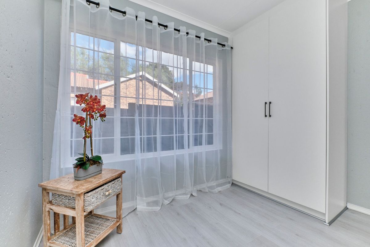 A bright, neutral bedroom featuring modern laminate flooring, a large grid-pattern window, and a clean, white built-in wardrobe.