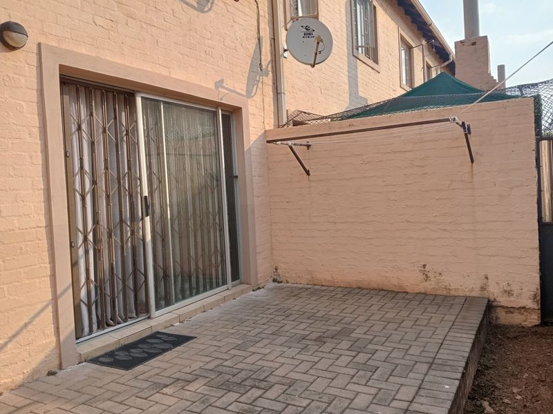 A paved rear patio area featuring a sliding glass door with security gate, wall-mounted clothesline, and exterior brick walling.