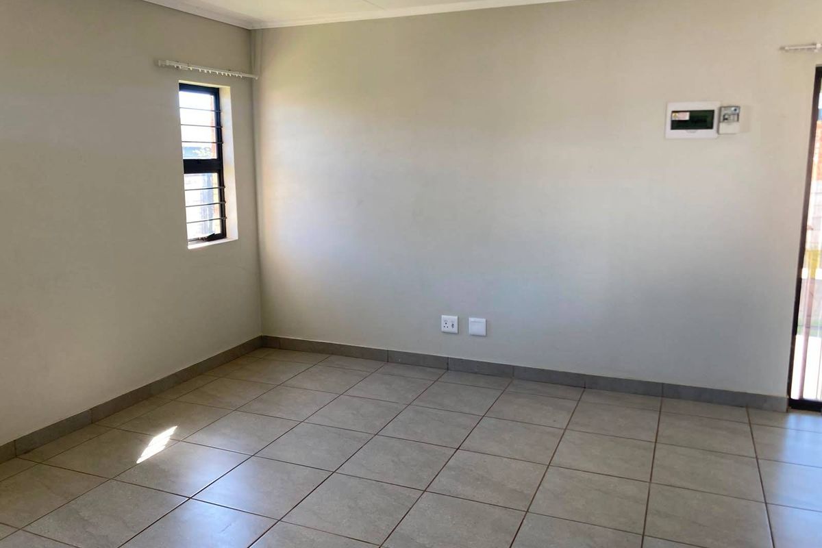 A clean, empty living area featuring neutral tiled flooring, recessed lighting, and a window with security bars.