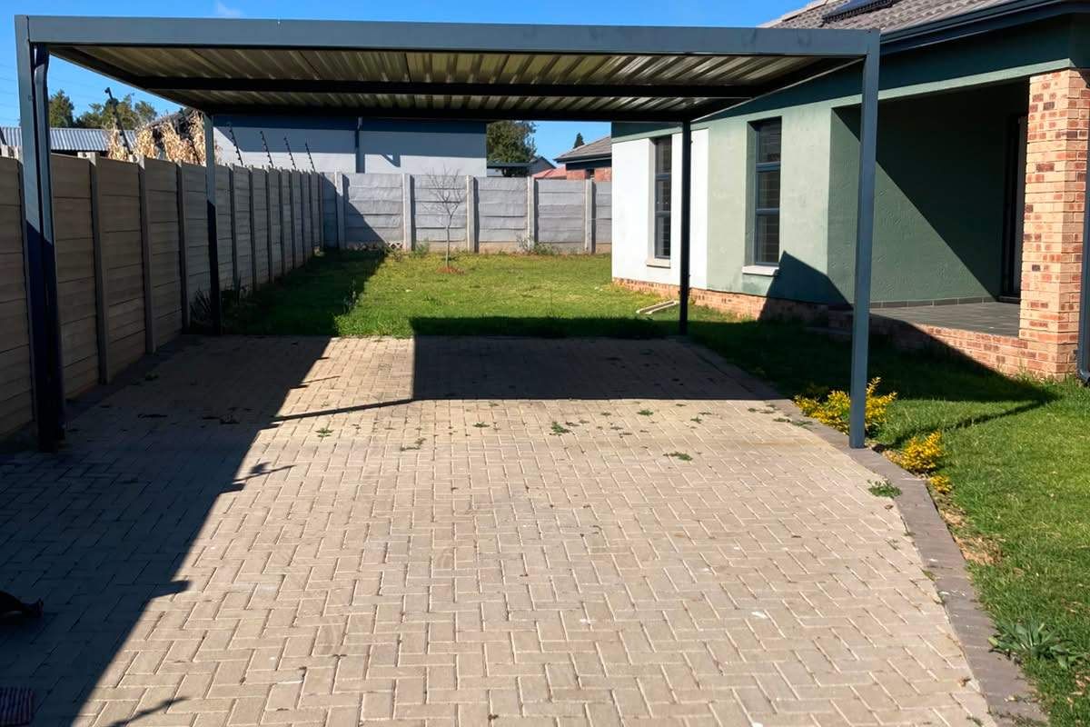 Rear exterior view featuring a paved driveway with a metal carport structure, precast boundary wall, and solar water heating system on the roof.