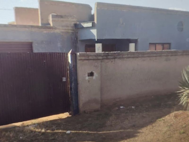 Front exterior view of a single-story house featuring a perimeter wall, metal gate, and garage access, showing signs of significant weathering and maintenance needs.