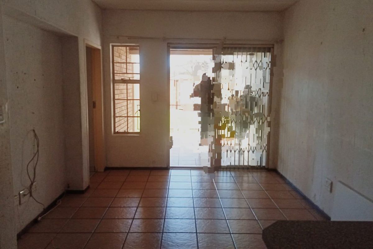 An empty living area featuring tiled flooring and a sliding security gate leading to the exterior, requiring cosmetic updates and wall repairs.