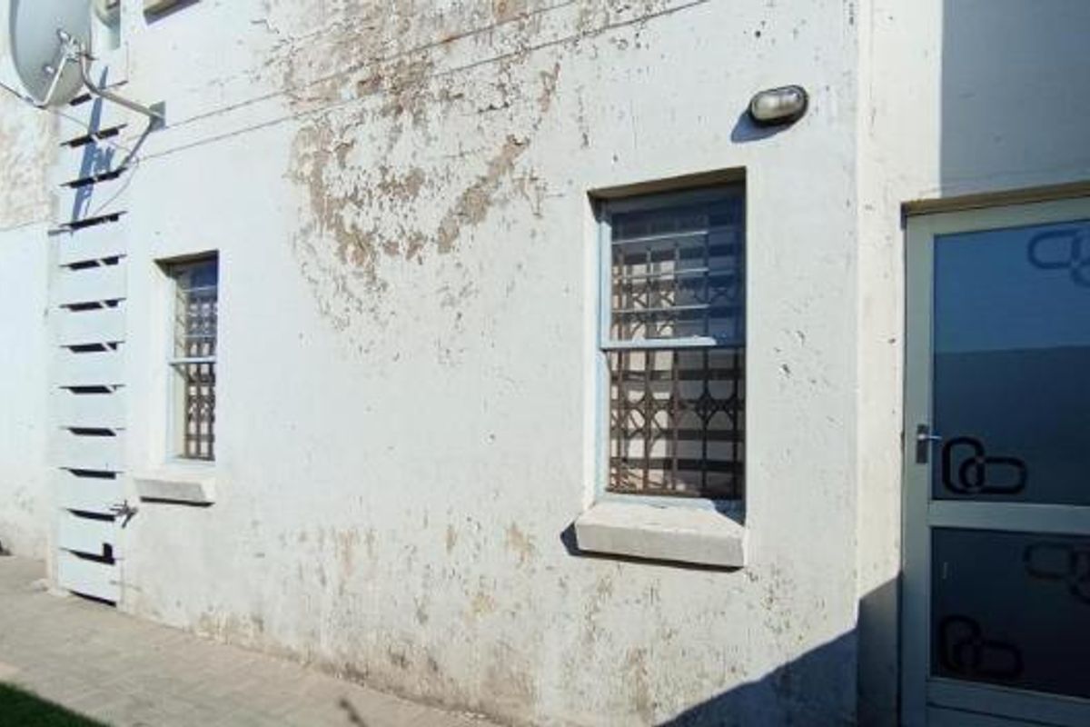 Rear exterior view of the apartment building showing paved walkway, security bars on windows, and weathered wall finishes requiring maintenance.