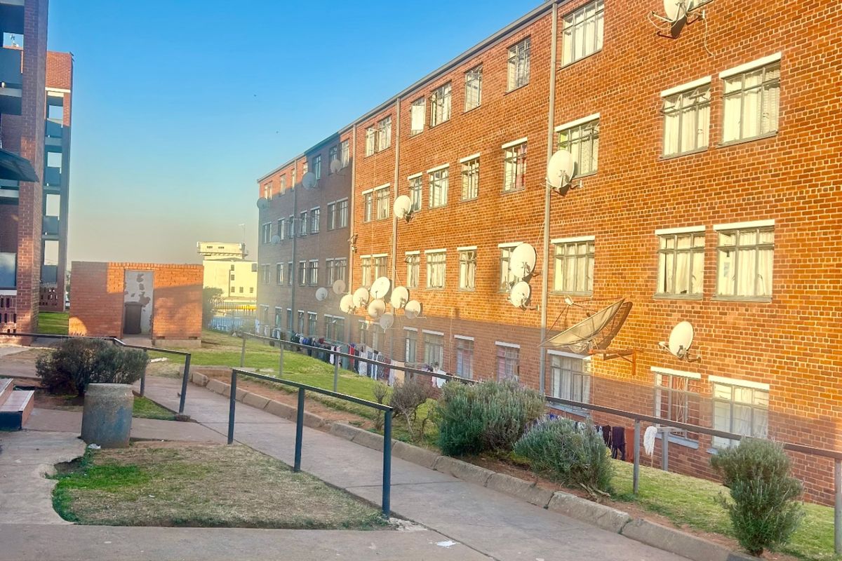 Exterior view of a multi-story brick apartment building featuring communal walkways, landscaped garden areas, and multiple satellite dish installations on the facade.