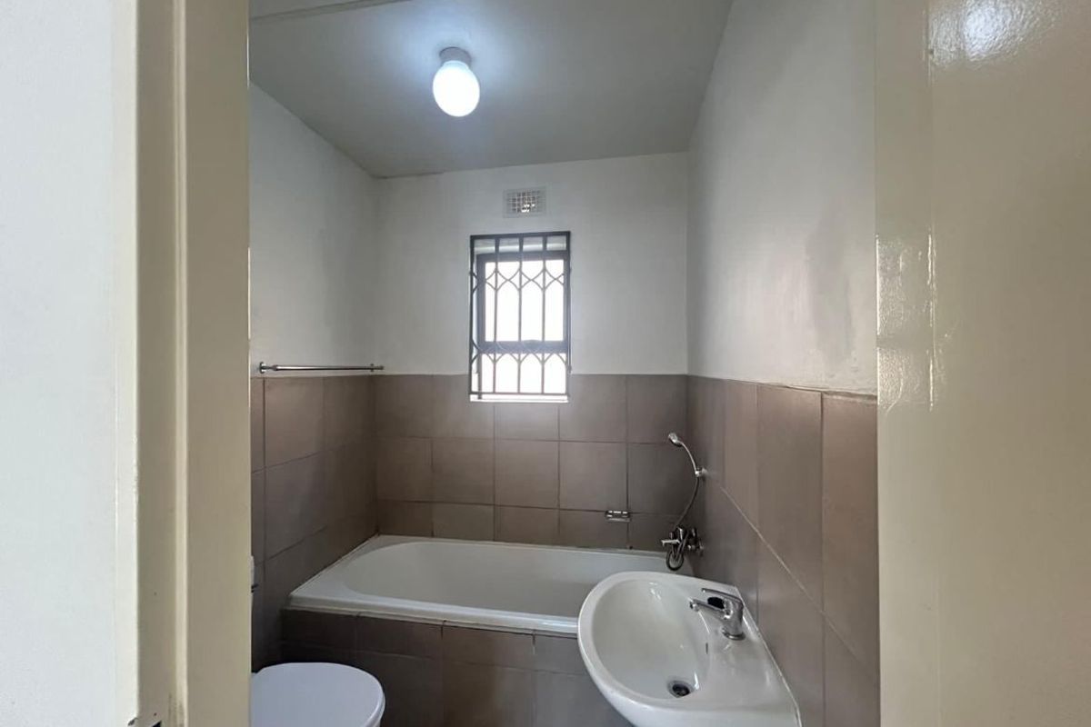 A functional bathroom featuring a bathtub, pedestal basin, toilet, and neutral wall tiling with security bars on the window.