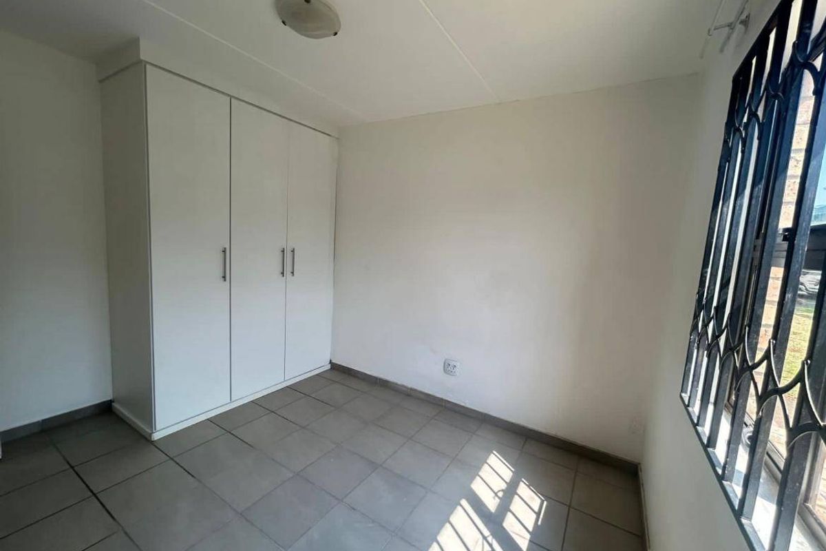 A clean, empty bedroom featuring neutral tiled flooring, a built-in wardrobe, and a window fitted with security bars.